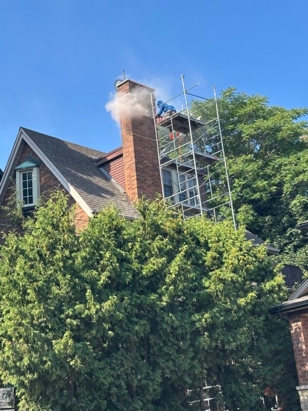 Brick chimney with scaffolding beside a pitched roof and mature garden trees
