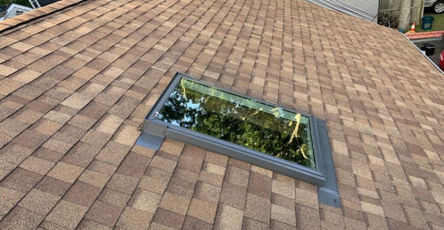 Glazed skylight set into a tan shingle roof, reflecting nearby tree branches