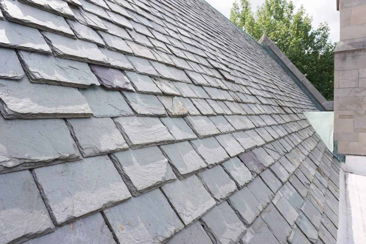 Grey slate roof slope running beside a stone parapet wall with green trees behind