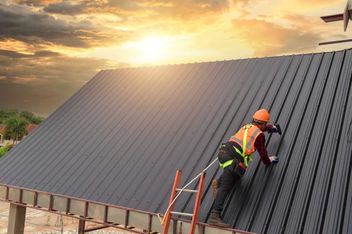 Installer in a high-visibility vest fixing dark metal roof sheets at sunset